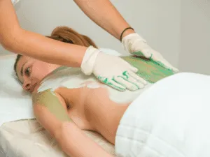 Estetista che applica fango per il corpo verde sulla schiena di una cliente come trattamento di fanghi anticellulite e relax.