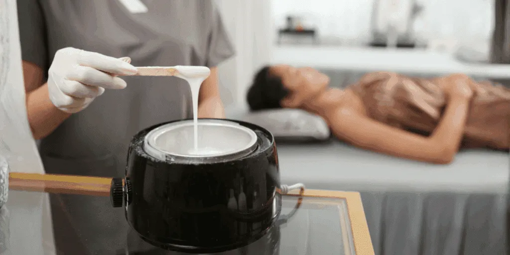Estetista che prepara la ceretta a caldo con un bastoncino, pronta per essere applicata sulla pelle di una cliente sdraiata.