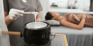 Estetista che prepara la ceretta a caldo con un bastoncino, pronta per essere applicata sulla pelle di una cliente sdraiata.