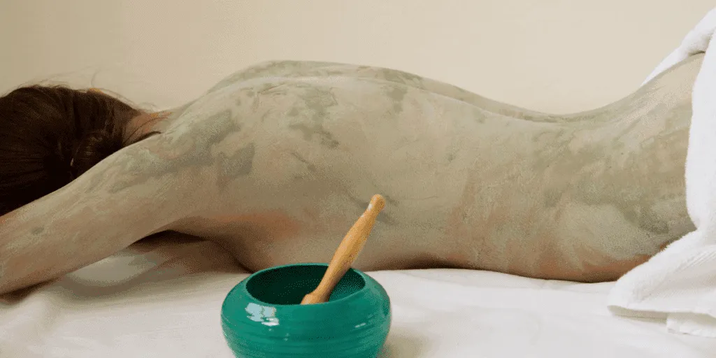 Donna distesa con il corpo ricoperto di fango per il corpo durante un trattamento di fanghi anticellulite e fanghi riducenti in centro estetico.