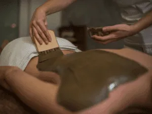 Operatrice estetica mentre applica fango per il corpo sulla schiena di una cliente durante un trattamento di fanghi anticellulite e fanghi riducenti.