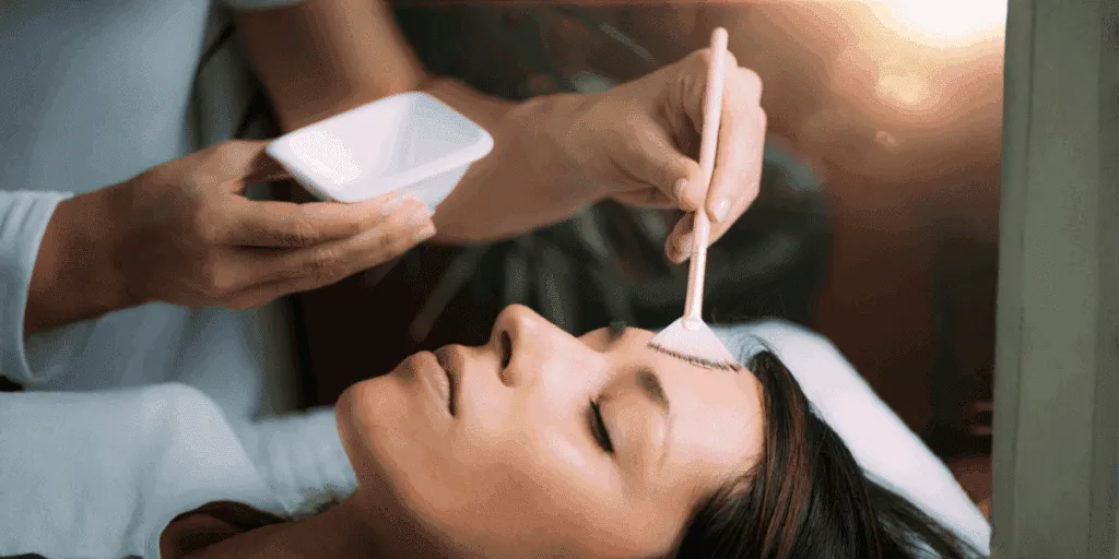 Estetista che applica un peeling enzimatico sul viso di una donna distesa durante un trattamento professionale.