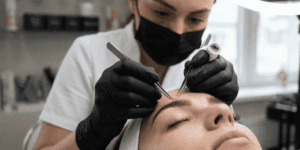 Dettaglio macro del trattamento di extension sopracciglia professionale: l'operatrice applica singole fibre sintetiche con pinzette di precisione per un effetto volume 3D naturale.
