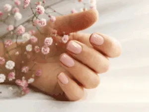 Unghie corte con smalto nude dopo manicure estetica professionale, fotografate accanto a piccoli fiori rosa.